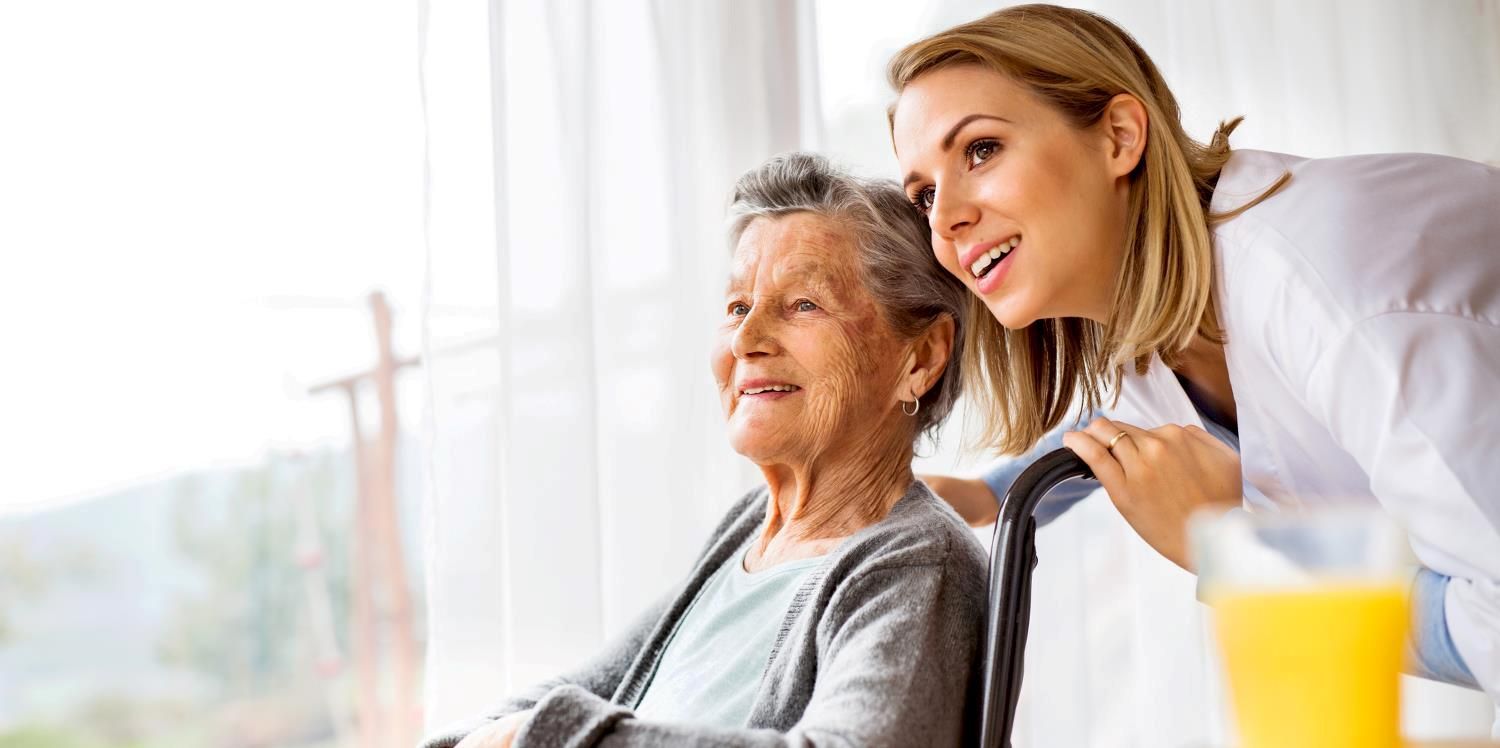 Elderly woman with wheelchair and caregiver smiling, looking outside